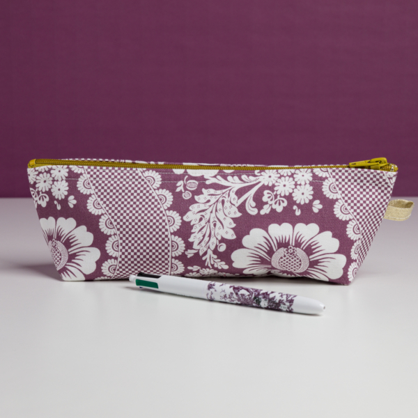 La XCOMM_TROUSSE STYLOS FLEURS AMARANTE est une pochette zippée à motif floral blanc et violet, présentée sur une surface blanche avec un stylo assorti devant, sur un fond violet uni. la marque Toile de Jouy