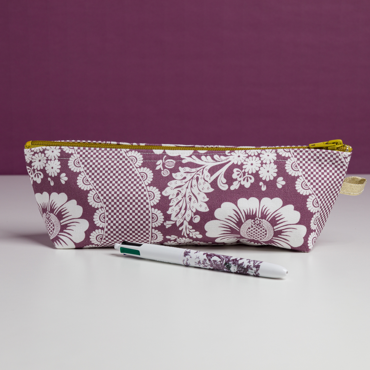 La XCOMM_TROUSSE STYLOS FLEURS AMARANTE est une pochette zippée à motif floral blanc et violet, présentée sur une surface blanche avec un stylo assorti devant, sur un fond violet uni. la marque Toile de Jouy