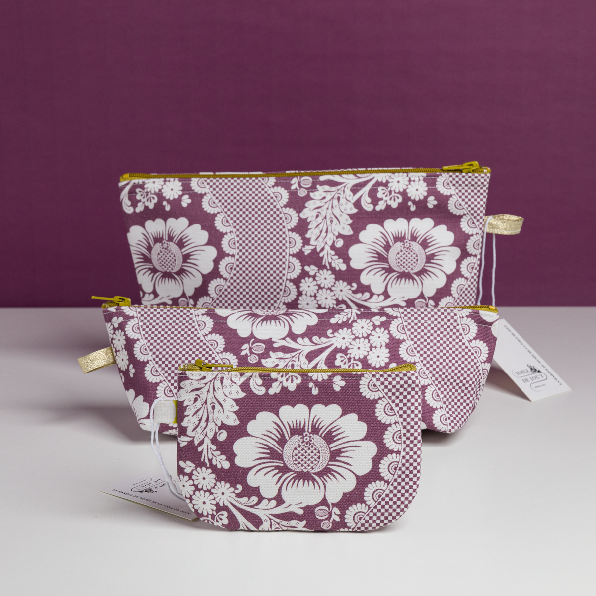 Trois pochettes XCOMM_TROUSSE STYLOS FLEURS AMARANTE avec des motifs floraux blancs et des fermetures éclair dorées sont disposées du plus petit au plus grand sur un fond violet, chacune comportant une étiquette blanche. la marque Toile de Jouy