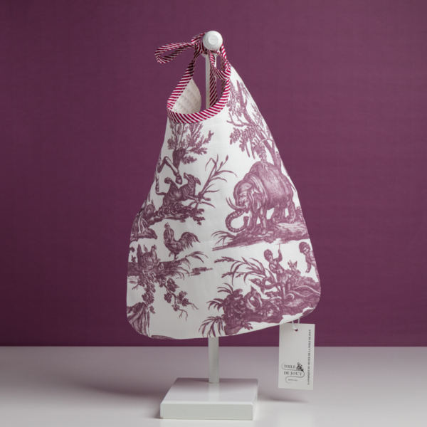 Le modèle XCOMM_BAVOIR 4 APRTIES AMARANTE est un sac en tissu blanc orné d'un imprimé violet représentant des animaux et des paysages, agrémenté d'une poignée en ruban et d'une étiquette, présenté sur un présentoir blanc devant un fond violet. la marque Toile de Jouy