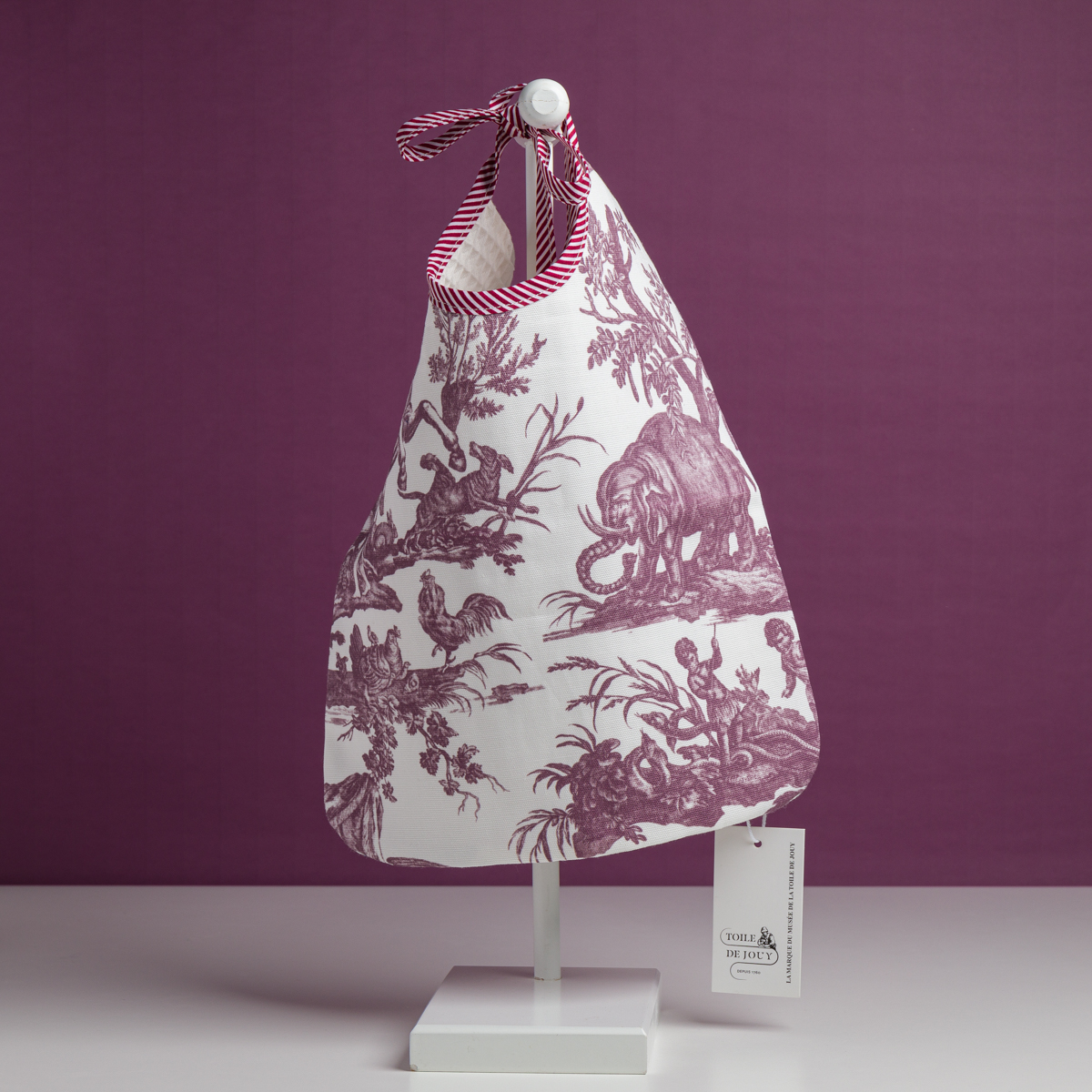 Le modèle XCOMM_BAVOIR 4 APRTIES AMARANTE est un sac en tissu blanc orné d'un imprimé violet représentant des animaux et des paysages, agrémenté d'une poignée en ruban et d'une étiquette, présenté sur un présentoir blanc devant un fond violet. la marque Toile de Jouy