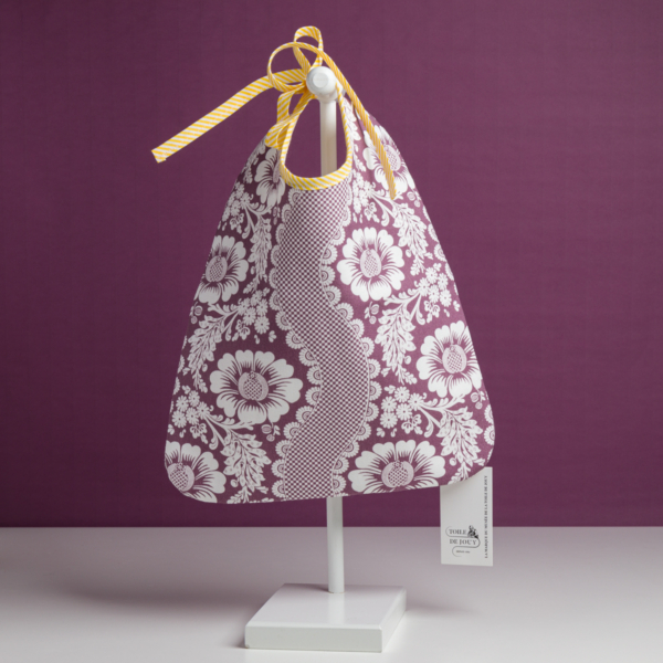 Le modèle XCOMM_BAVOIR 4 APRTIES AMARANTE, orné d'un motif floral violet et blanc, est présenté sur un présentoir blanc orné d'un ruban jaune noué en haut, sur fond violet. Une petite étiquette est accrochée dans le coin inférieur droit du sac. la marque Toile de Jouy