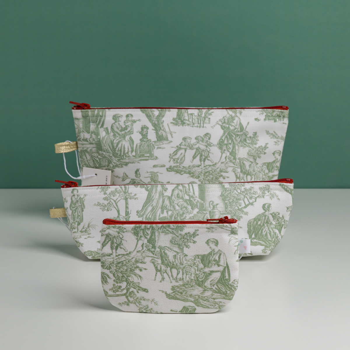 Trois pochettes XCOMM_TROUSSE STYLOS OFFRANDE SAUGE, en tissu blanc orné d'illustrations pastorales vertes et dotées de fermetures éclair rouges, sont disposées sur une surface blanche devant un fond vert, dans les tailles petite, moyenne et grande. la marque Toile de Jouy