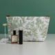 La trousse de toilette XCOMM_TROUSSE STYLOS OFFRANDE SAUGE, ornée d'illustrations pastorales vertes, repose sur une surface blanche. Devant elle se trouvent un flacon de parfum rose et un tube de rouge à lèvres noir, tandis qu'une étiquette blanche nouée d'un ruban doré est posée à côté de la trousse. L'arrière-plan est vert. la marque Toile de Jouy