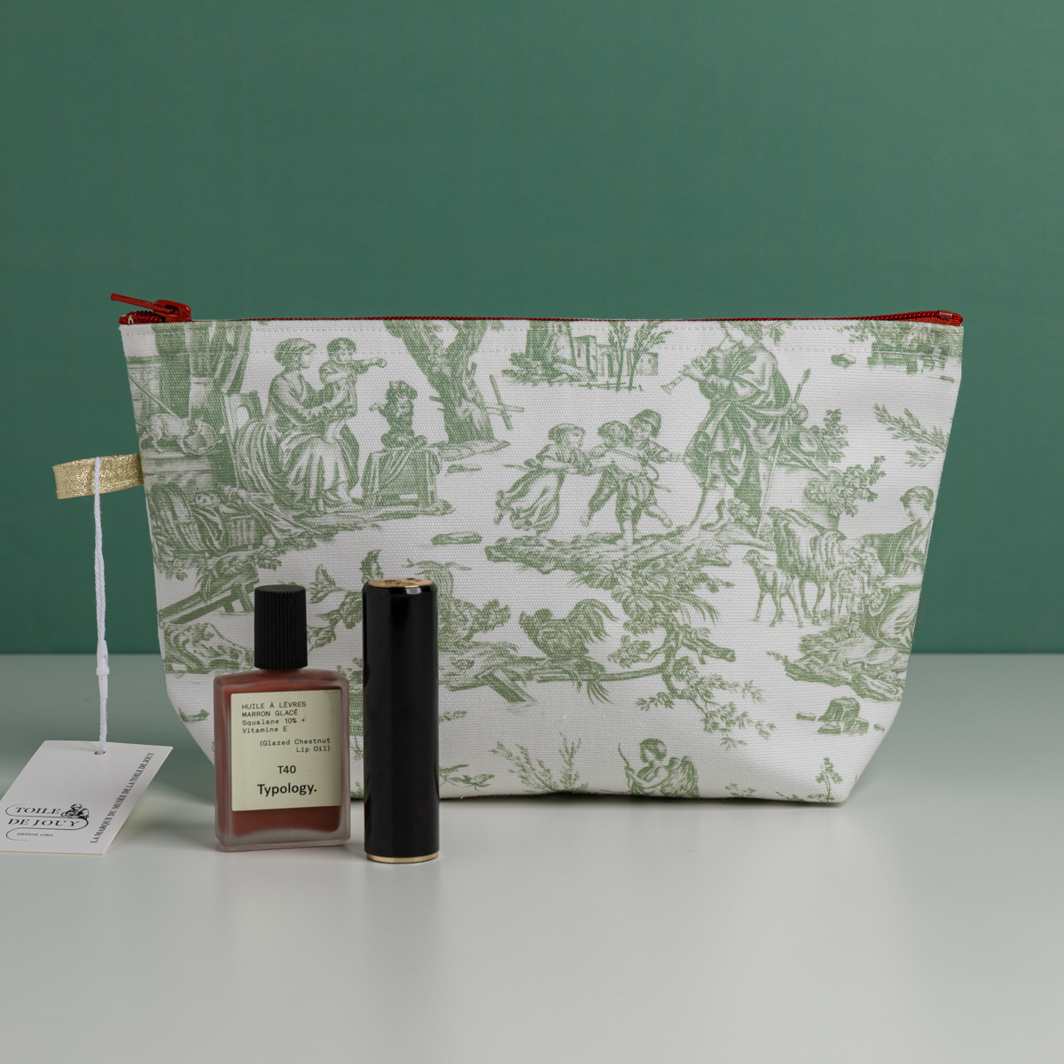 La trousse de toilette XCOMM_TROUSSE STYLOS OFFRANDE SAUGE, ornée d'illustrations pastorales vertes, repose sur une surface blanche. Devant elle se trouvent un flacon de parfum rose et un tube de rouge à lèvres noir, tandis qu'une étiquette blanche nouée d'un ruban doré est posée à côté de la trousse. L'arrière-plan est vert. la marque Toile de Jouy