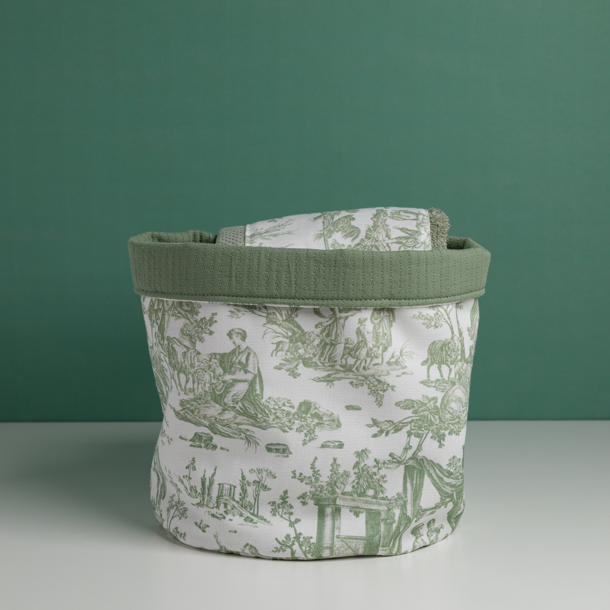 Le XCOMM_CACHE POT-CORBEILE OFFRANDE SAUGE, un panier de rangement en tissu avec un bord vert et un imprimé pastoral vert et blanc, est posé sur une surface blanche sur fond vert avec un tissu plié assorti à l'intérieur. la marque Toile de Jouy
