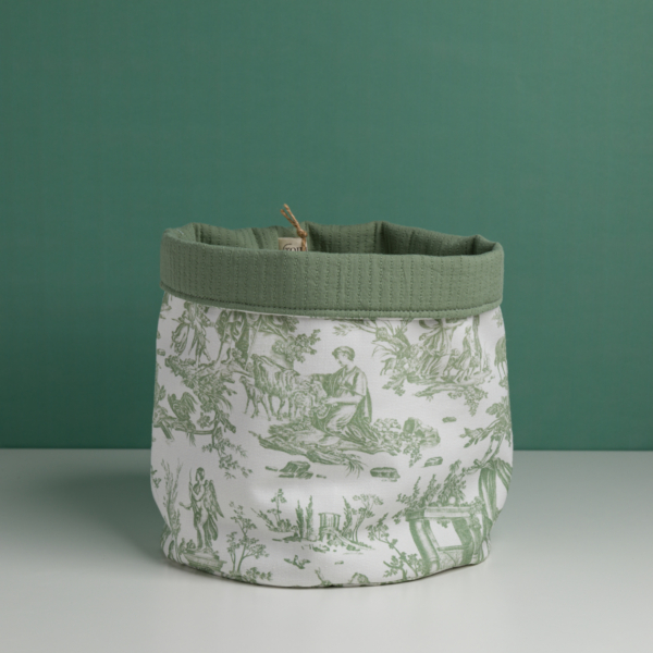 Le XCOMM_CACHE POT-CORBEILE OFFRANDE SAUGE, un panier de rangement en tissu imprimé d'une toile verte représentant des scènes pastorales de personnes et d'animaux et terminé par un rebord vert, est présenté sur une surface blanche sur fond vert. la marque Toile de Jouy