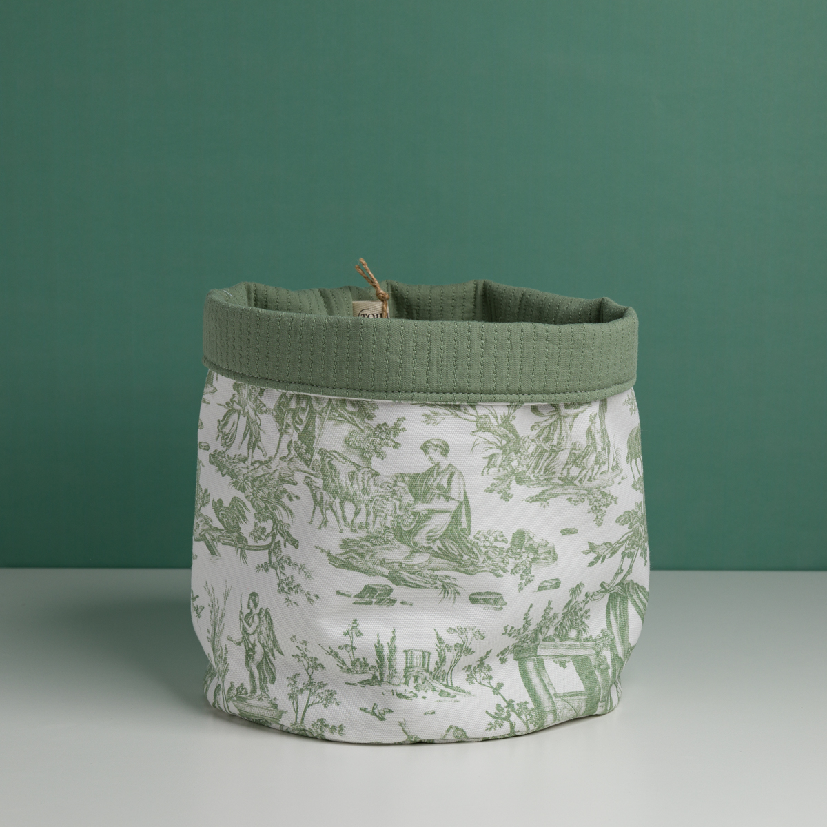 Le XCOMM_CACHE POT-CORBEILE OFFRANDE SAUGE, un panier de rangement en tissu imprimé d'une toile verte représentant des scènes pastorales de personnes et d'animaux et terminé par un rebord vert, est présenté sur une surface blanche sur fond vert. la marque Toile de Jouy