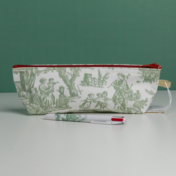 Une pochette blanche « XCOMM_TROUSSE STYLOS OFFRANDE SAUGE », ornée de scènes pastorales vertes, est posée à la verticale sur une surface blanche. Devant elle repose un stylo blanc assorti, arborant les mêmes motifs verts. L'arrière-plan est d'un vert uni. la marque Toile de Jouy