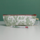 Une pochette blanche « XCOMM_TROUSSE STYLOS OFFRANDE SAUGE », ornée de scènes pastorales vertes, est posée à la verticale sur une surface blanche. Devant elle repose un stylo blanc assorti, arborant les mêmes motifs verts. L'arrière-plan est d'un vert uni. la marque Toile de Jouy