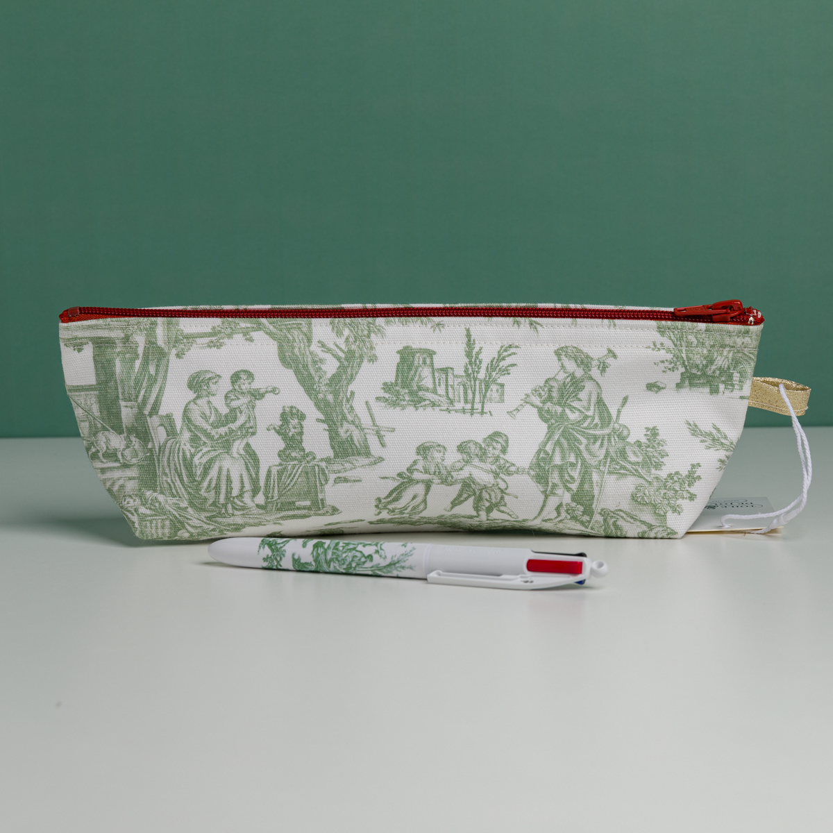 Une pochette blanche « XCOMM_TROUSSE STYLOS OFFRANDE SAUGE », ornée de scènes pastorales vertes, est posée à la verticale sur une surface blanche. Devant elle repose un stylo blanc assorti, arborant les mêmes motifs verts. L'arrière-plan est d'un vert uni. la marque Toile de Jouy
