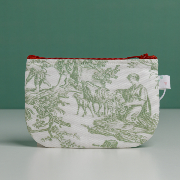 La « XCOMM_TROUSSE STYLOS OFFRANDE SAUGE » est une petite pochette en tissu dotée d'une fermeture éclair rouge, ornée d'un imprimé représentant une scène champêtre verte avec des personnages et des animaux. Elle est posée à la verticale sur une surface unie, sur fond vert. la marque Toile de Jouy