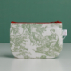 La « XCOMM_TROUSSE STYLOS OFFRANDE SAUGE » est une petite pochette en tissu dotée d'une fermeture éclair rouge, ornée d'un imprimé représentant une scène champêtre verte avec des personnages et des animaux. Elle est posée à la verticale sur une surface unie, sur fond vert. la marque Toile de Jouy