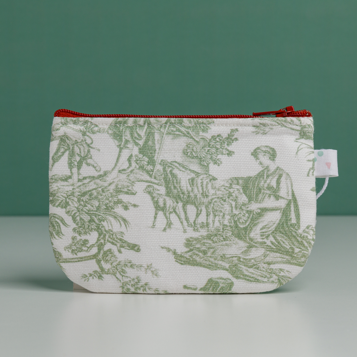 La « XCOMM_TROUSSE STYLOS OFFRANDE SAUGE » est une petite pochette en tissu dotée d'une fermeture éclair rouge, ornée d'un imprimé représentant une scène champêtre verte avec des personnages et des animaux. Elle est posée à la verticale sur une surface unie, sur fond vert. la marque Toile de Jouy