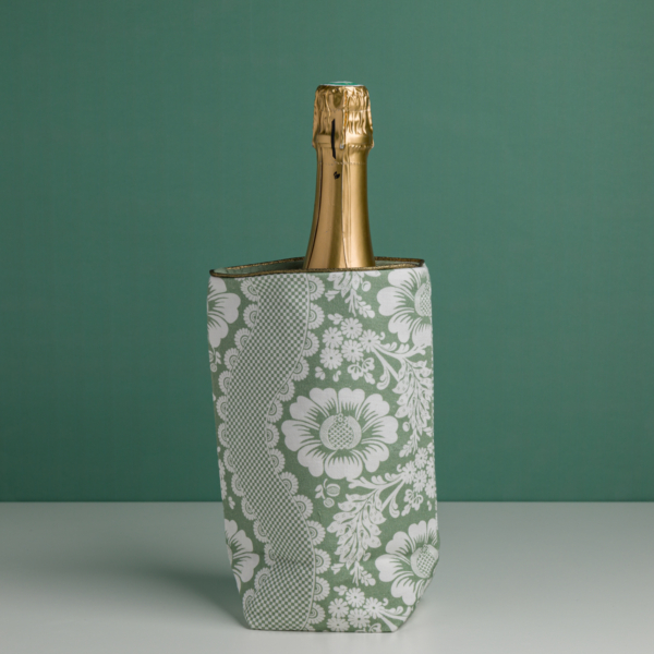 Le XCOMM_HOUSSE BOUTEILLE OFFRANDE SAUGE, un rafraîchisseur floral vert et blanc, contient une bouteille de champagne à bouchon doré sur un fond vert assorti. la marque Toile de Jouy