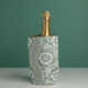 Le XCOMM_HOUSSE BOUTEILLE OFFRANDE SAUGE, un rafraîchisseur floral vert et blanc, contient une bouteille de champagne à bouchon doré sur un fond vert assorti. la marque Toile de Jouy