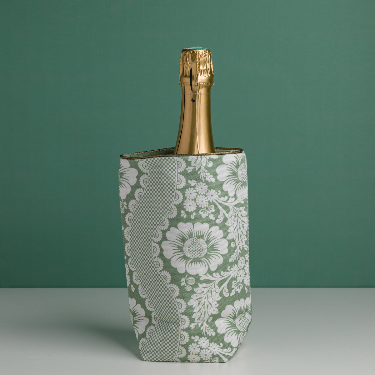 Le XCOMM_HOUSSE BOUTEILLE OFFRANDE SAUGE, un rafraîchisseur floral vert et blanc, contient une bouteille de champagne à bouchon doré sur un fond vert assorti. la marque Toile de Jouy