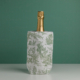 La bouteille de champagne XCOMM_HOUSSE BOUTEILLE OFFRANDE SAUGE, recouverte d'une feuille d'or, est placée dans un refroidisseur à vin décoratif à motifs blancs et verts sur fond vert uni. la marque Toile de Jouy