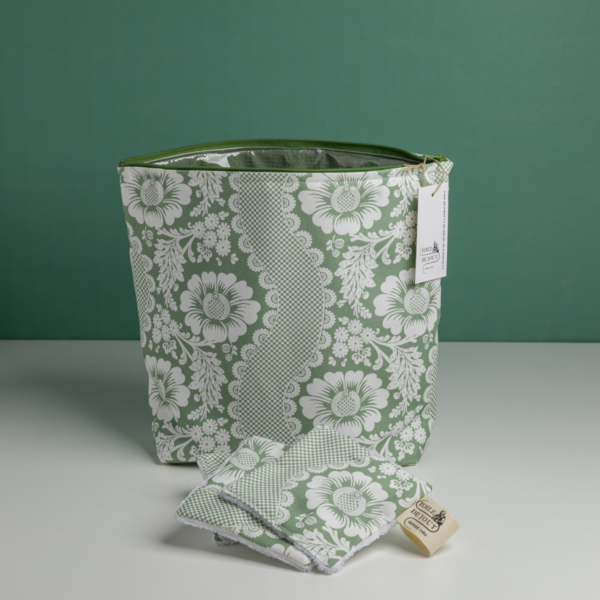 La « XCOMM_GRANDE TROUSSE DE TOILETTE FLEURS AMARANTE », une trousse en tissu à motifs floraux verts et blancs munie d'une étiquette, est posée sur une surface blanche, avec des lingettes réutilisables assorties pliées devant elle, sur fond vert. la marque Toile de Jouy