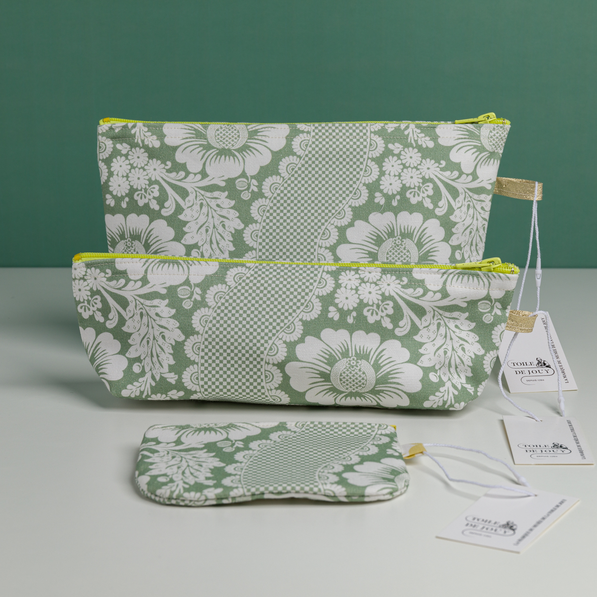 Deux pochettes XCOMM_TROUSSE STYLOS FLEURS SAUGE et un petit porte-monnaie, tous avec des motifs floraux et dentelés blancs, sont présentés sur une surface blanche sur fond vert, chacun avec une étiquette blanche. la marque Toile de Jouy