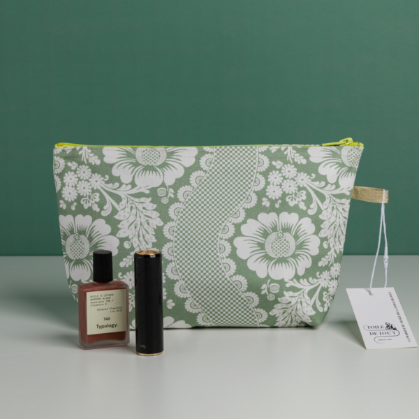 Une XCOMM_TROUSSE STYLOS FLEURS SAUGE, une pochette à fermeture éclair à motif floral vert et blanc, est placée sur une surface blanche avec un vernis à ongles Typology, un tube de rouge à lèvres noir et une étiquette blanche à l'avant. Le fond est vert. la marque Toile de Jouy