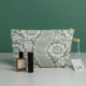 Une XCOMM_TROUSSE STYLOS FLEURS SAUGE, une pochette à fermeture éclair à motif floral vert et blanc, est placée sur une surface blanche avec un vernis à ongles Typology, un tube de rouge à lèvres noir et une étiquette blanche à l'avant. Le fond est vert. la marque Toile de Jouy