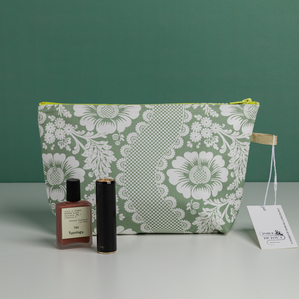 Une XCOMM_TROUSSE STYLOS FLEURS SAUGE, une pochette à fermeture éclair à motif floral vert et blanc, est placée sur une surface blanche avec un vernis à ongles Typology, un tube de rouge à lèvres noir et une étiquette blanche à l'avant. Le fond est vert. la marque Toile de Jouy