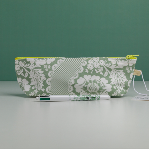 La XCOMM_TROUSSE STYLOS FLEURS SAUGE, une trousse à crayons à motifs floraux verts et blancs avec une fermeture éclair jaune, repose sur une surface claire. Devant, un stylo blanc assorti est orné de motifs floraux verts sur un fond vert uni. la marque Toile de Jouy