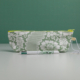 La XCOMM_TROUSSE STYLOS FLEURS SAUGE, une trousse à crayons à motifs floraux verts et blancs avec une fermeture éclair jaune, repose sur une surface claire. Devant, un stylo blanc assorti est orné de motifs floraux verts sur un fond vert uni. la marque Toile de Jouy