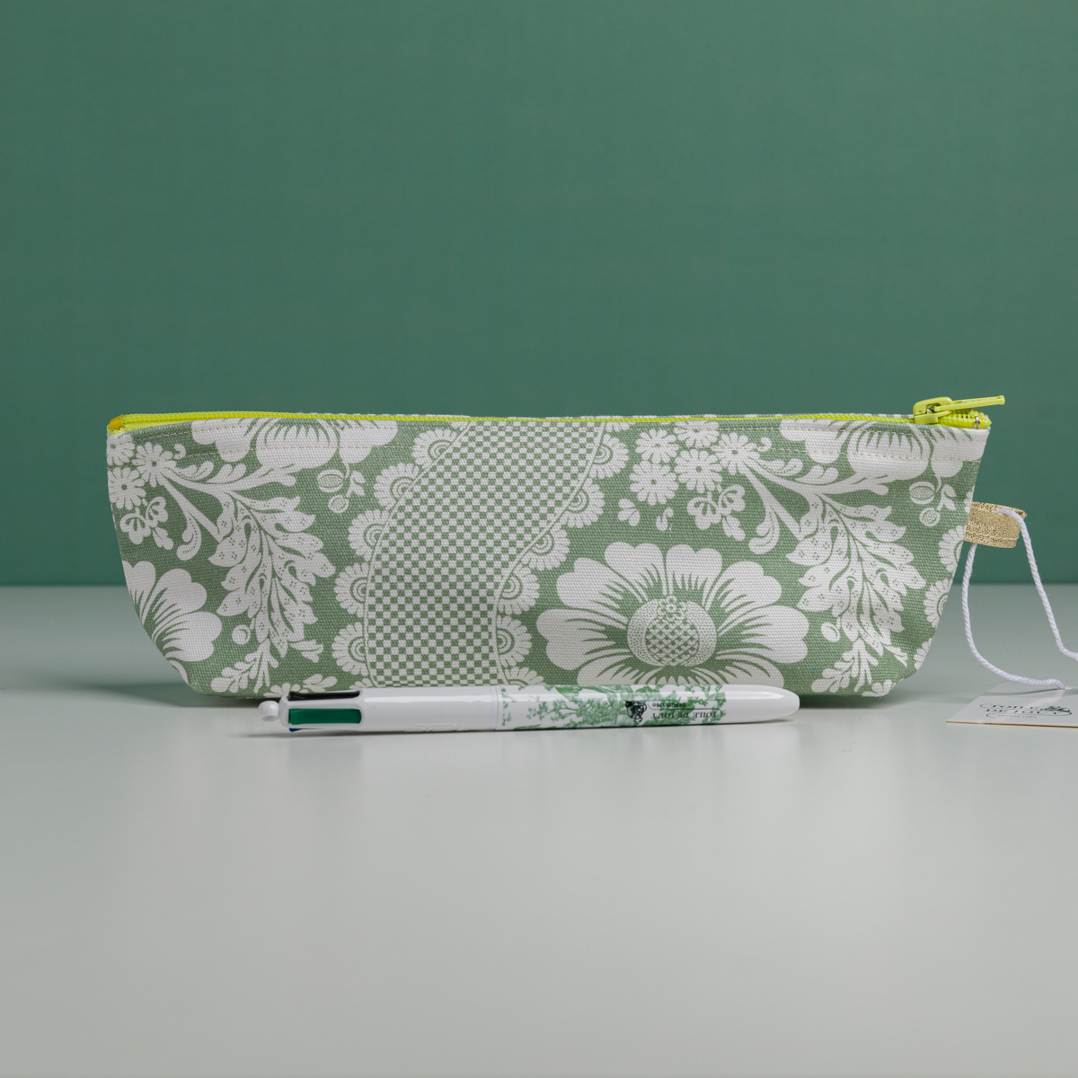 La XCOMM_TROUSSE STYLOS FLEURS SAUGE, une trousse à crayons à motifs floraux verts et blancs avec une fermeture éclair jaune, repose sur une surface claire. Devant, un stylo blanc assorti est orné de motifs floraux verts sur un fond vert uni. la marque Toile de Jouy