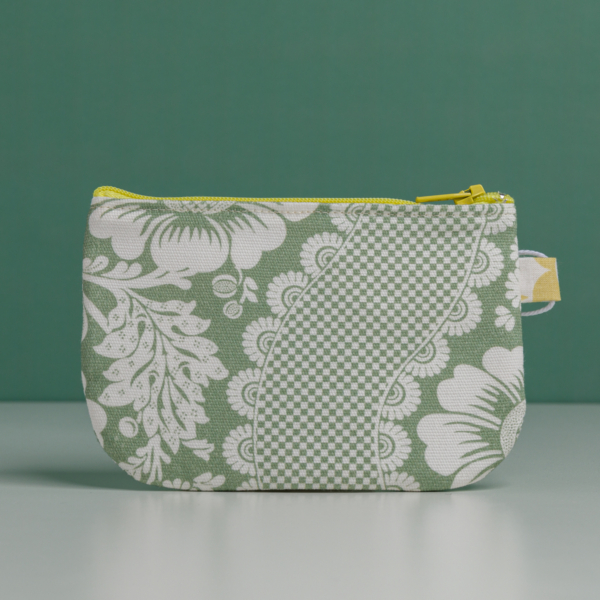 La XCOMM_TROUSSE STYLOS FLEURS SAUGE est une petite pochette à fermeture éclair avec un motif floral à carreaux verts et blancs, avec une fermeture éclair jaune et une étiquette jaune sur le côté droit, présentée à la verticale sur un fond vert uni. la marque Toile de Jouy