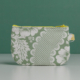 La XCOMM_TROUSSE STYLOS FLEURS SAUGE est une petite pochette à fermeture éclair avec un motif floral à carreaux verts et blancs, avec une fermeture éclair jaune et une étiquette jaune sur le côté droit, présentée à la verticale sur un fond vert uni. la marque Toile de Jouy