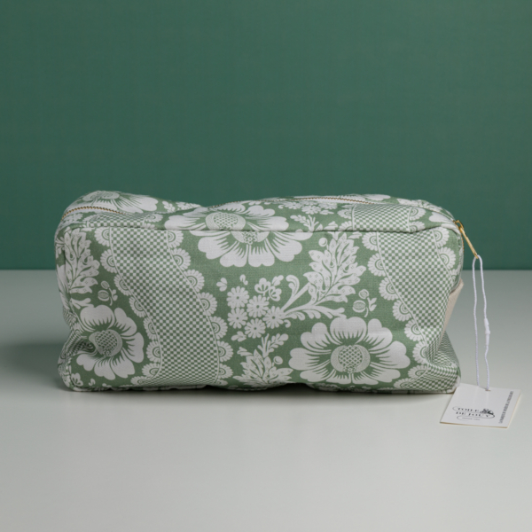 La « XCOMM_TROUSSE RECTANGULAIRE FLEUS SAUGE » est une pochette en tissu vert ornée d'un motif floral blanc, dotée d'une fermeture à glissière et d'une étiquette sur la tirette. Elle est photographiée sur une surface blanche, sur fond vert. la marque Toile de Jouy