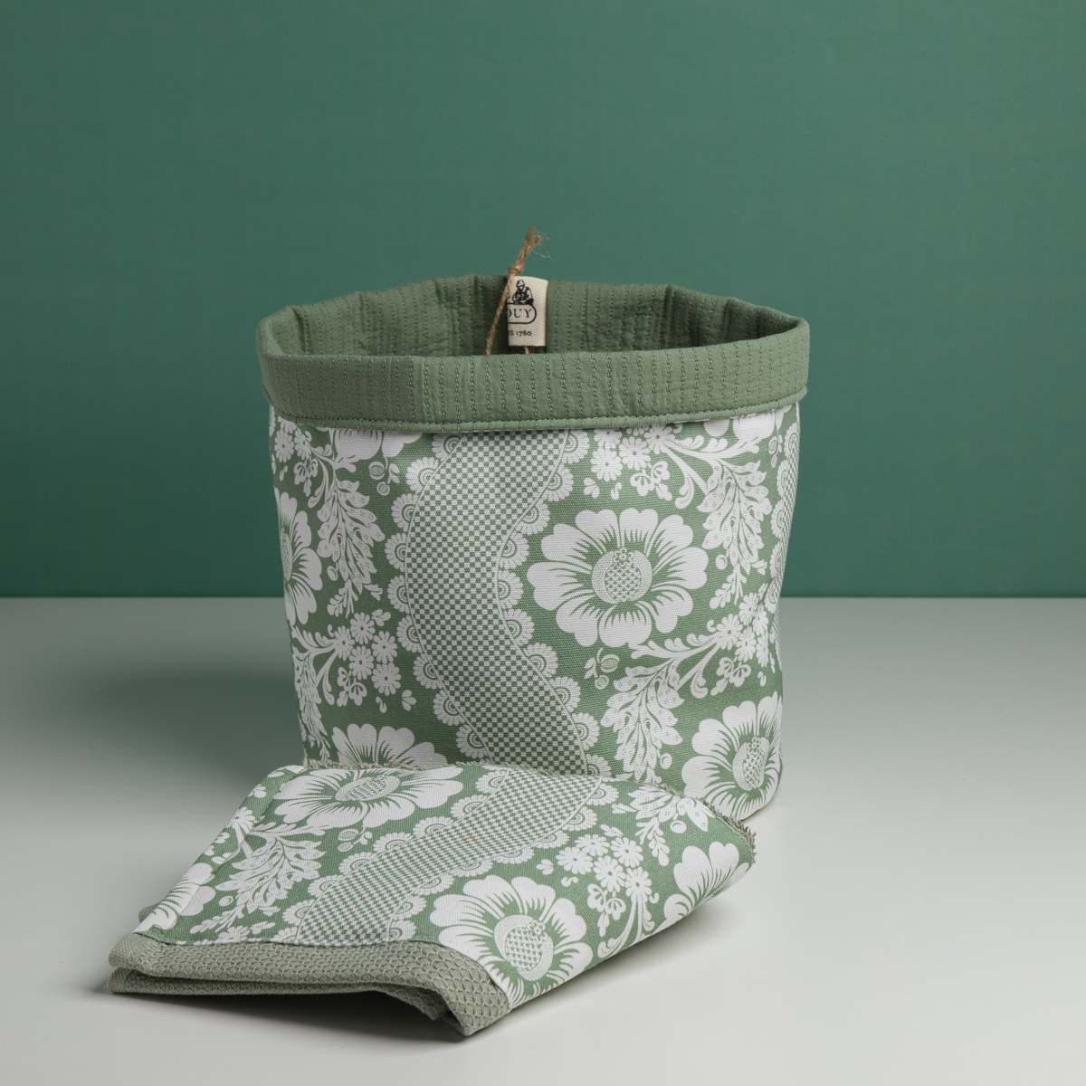 Le XCOMM_CACHE POT-CORBEILE OFFRANDE SAUGE, un panier de rangement en tissu floral vert et blanc, est posé sur une surface blanche devant un mur vert assorti. Devant lui se trouve une serviette pliée avec le même motif floral. la marque Toile de Jouy