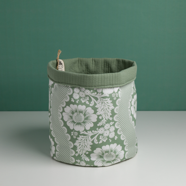 Le XCOMM_CACHE POT-CORBEILE OFFRANDE SAUGE est un panier de rangement en tissu vert avec un motif floral blanc, une doublure verte unie et une petite étiquette sur le bord. Il est présenté sur une surface blanche à fond vert. la marque Toile de Jouy