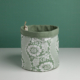 Le XCOMM_CACHE POT-CORBEILE OFFRANDE SAUGE est un panier de rangement en tissu vert avec un motif floral blanc, une doublure verte unie et une petite étiquette sur le bord. Il est présenté sur une surface blanche à fond vert. la marque Toile de Jouy