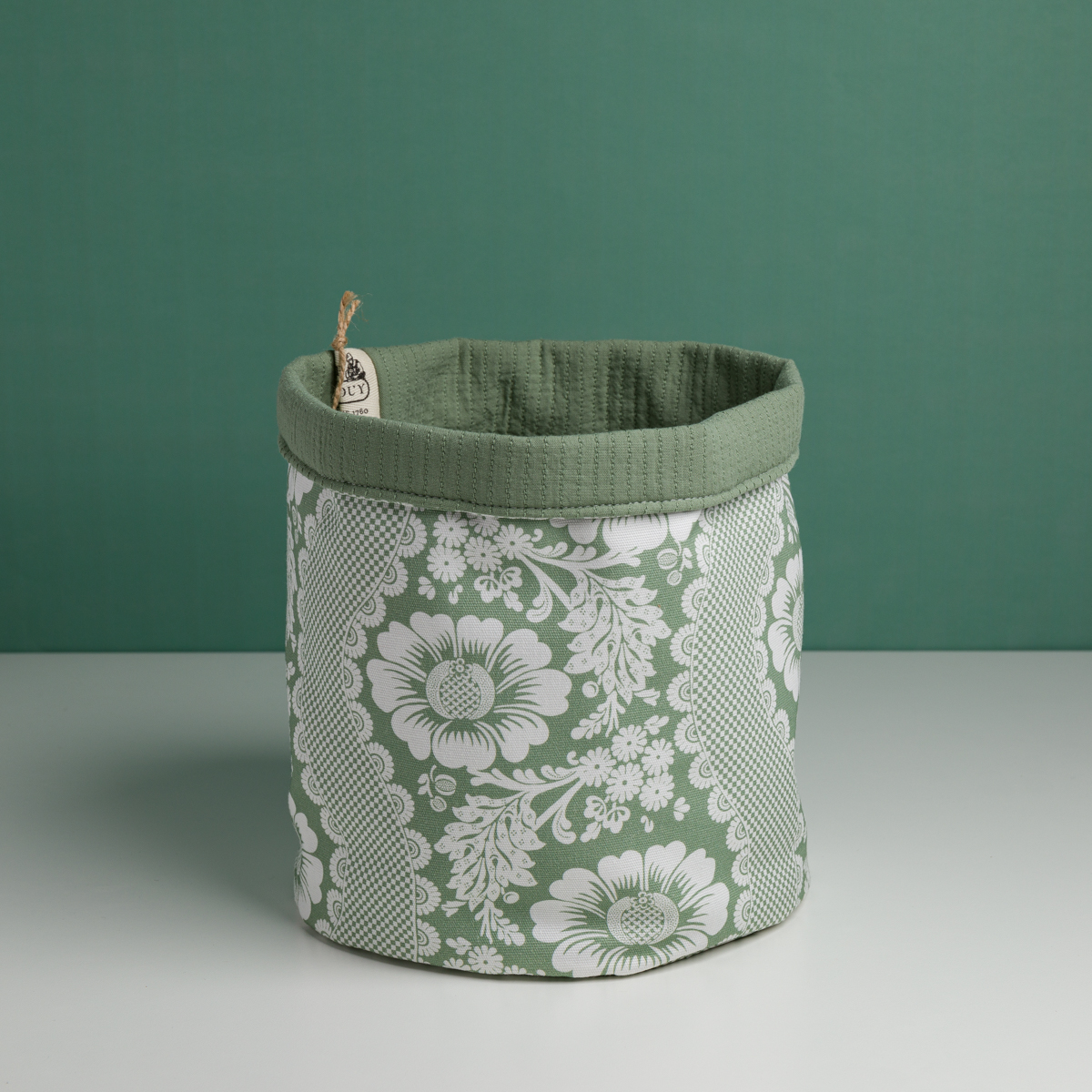 Le XCOMM_CACHE POT-CORBEILE OFFRANDE SAUGE est un panier de rangement en tissu vert avec un motif floral blanc, une doublure verte unie et une petite étiquette sur le bord. Il est présenté sur une surface blanche à fond vert. la marque Toile de Jouy