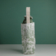 Le XCOMM_HOUSSE BOUTEILLE OFFRANDE SAUGE se tient debout sur une surface blanche sur fond vert, avec un imprimé pastoral blanc et vert, une poignée beige, et est rempli de tissu de couleur sauge. la marque Toile de Jouy