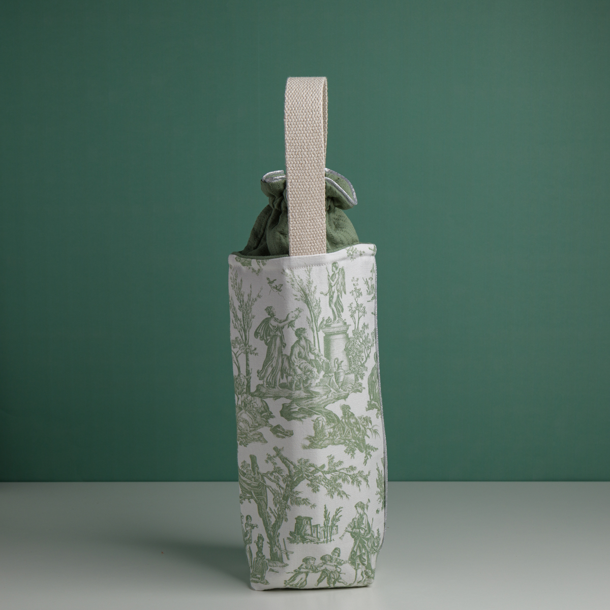 Le XCOMM_HOUSSE BOUTEILLE OFFRANDE SAUGE se tient debout sur une surface blanche sur fond vert, avec un imprimé pastoral blanc et vert, une poignée beige, et est rempli de tissu de couleur sauge. la marque Toile de Jouy