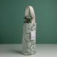 Un sac à vin XCOMM_HOUSSE BOUTEILLE OFFRANDE SAUGE présentant un motif floral vert et blanc avec une lanière beige se tient debout sur un fond vert assorti. Une étiquette "Cork & Be Field" est attachée au sac. la marque Toile de Jouy