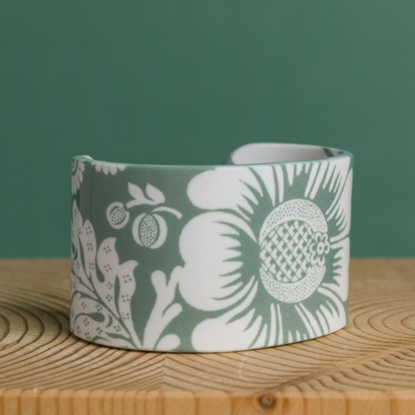La XCOMM_ESPADRILLE FLEURS MANDARINE P38, un bracelet en céramique blanche orné de motifs floraux et fruités verts, repose sur une surface en bois clair, sur fond vert. la marque Toile de Jouy