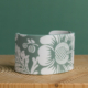 La XCOMM_ESPADRILLE FLEURS MANDARINE P38, un bracelet en céramique blanche orné de motifs floraux et fruités verts, repose sur une surface en bois clair, sur fond vert. la marque Toile de Jouy