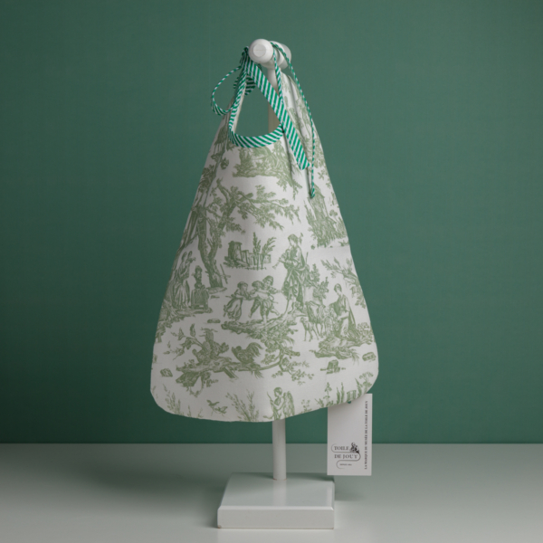 Le « XCOMM_TROUSSE STYLOS OFFRANDE SAUGE », un sac en tissu orné de scènes pastorales vertes et doté de poignées rayées vertes et blanches, est suspendu à un support blanc sur fond vert. Une étiquette comportant du texte est fixée au sac. la marque Toile de Jouy