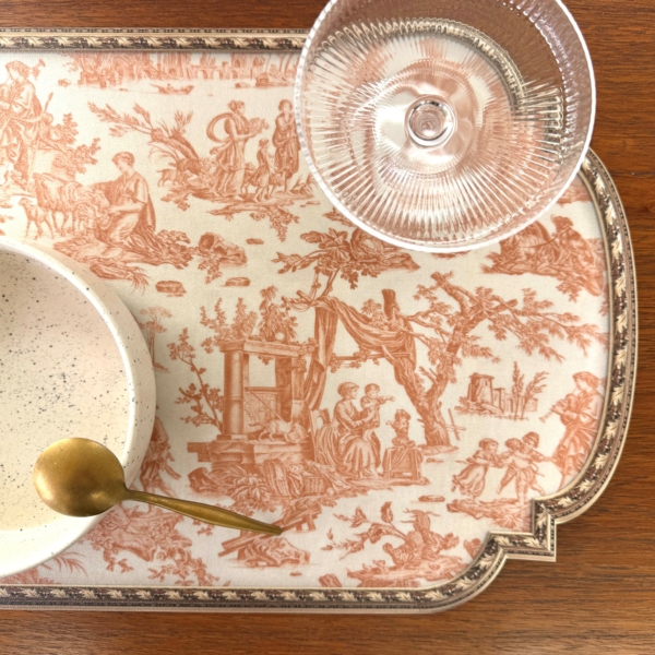 SAFRAN Un napperon décoratif avec des scènes complexes en brun-rouge, un bol blanc moucheté avec une cuillère en or et un gobelet en verre transparent sont posés sur une surface en bois. la marque Toile de Jouy