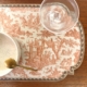 Le SET DE TABLE VINYLE "OFFRANDE A L'AMOUR" SAFRAN décore une table en bois, accompagné d'un plat en verre, d'un bol en céramique mouchetée et d'une cuillère en or. la marque Toile de Jouy