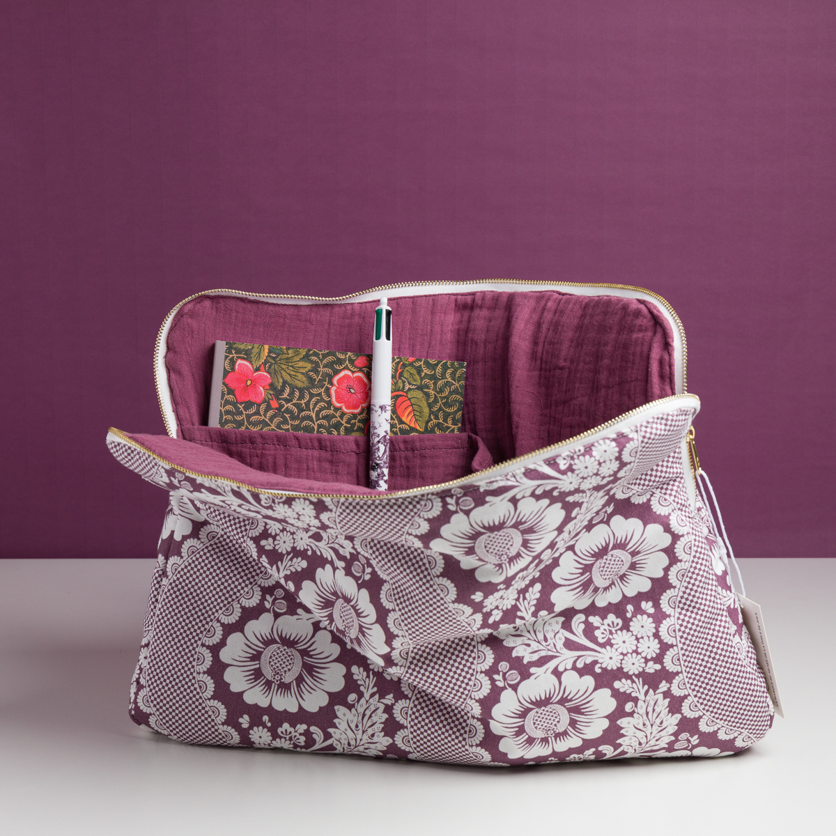 Un MATELAS DE REPOS "FLEURS STYLISéES" MANDARINE repose ouvert sur une surface blanche, montrant un stylo à fleurs et un carnet assorti à l'intérieur, avec un thème floral violet s'étendant à l'arrière-plan. la marque Toile de Jouy