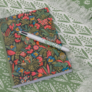 La MANDARINE MATELAS DE REPOS "FLEURS STYLISéES" présente un motif floral vibrant et repose sur un tissu à motifs verts et blancs avec des bords frangés. la marque Toile de Jouy