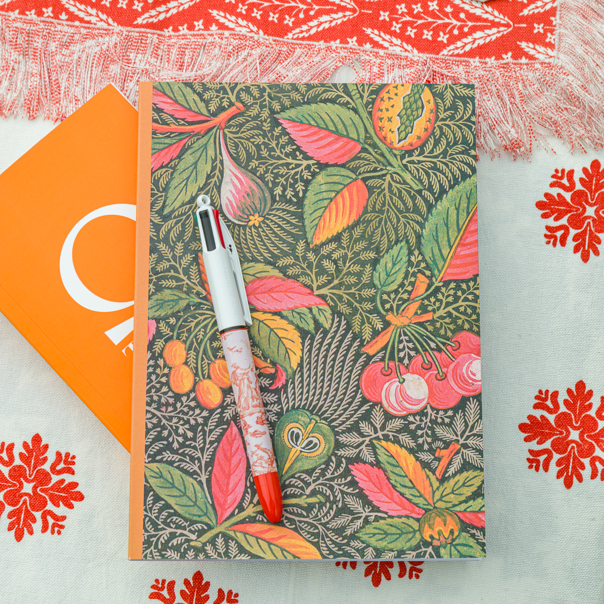 Le MATELAS DE REPOS "FLEURS STYLISéES" MANDARINE repose sur un tissu blanc brodé de fleurs rouges, aux côtés d'un carnet botanique coloré et d'un stylo rouge et blanc. la marque Toile de Jouy