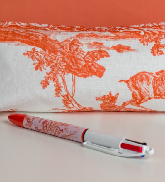Le MATELAS DE REPOS "FLEURS STYLISéES" MANDARINE, avec son motif floral orange, repose sur une surface blanche contre un mur orange, accompagné d'un stylo multicolore rouge et blanc au premier plan. la marque Toile de Jouy