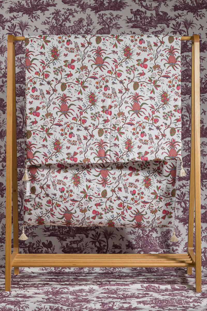 Le MATELAS DE REPOS "ANANAS", un support en bois contenant un tissu au motif floral rouge, vert et or, est placé sur un papier peint botanique et pastoral violet pour un design coordonné et superposé. la marque Toile de Jouy